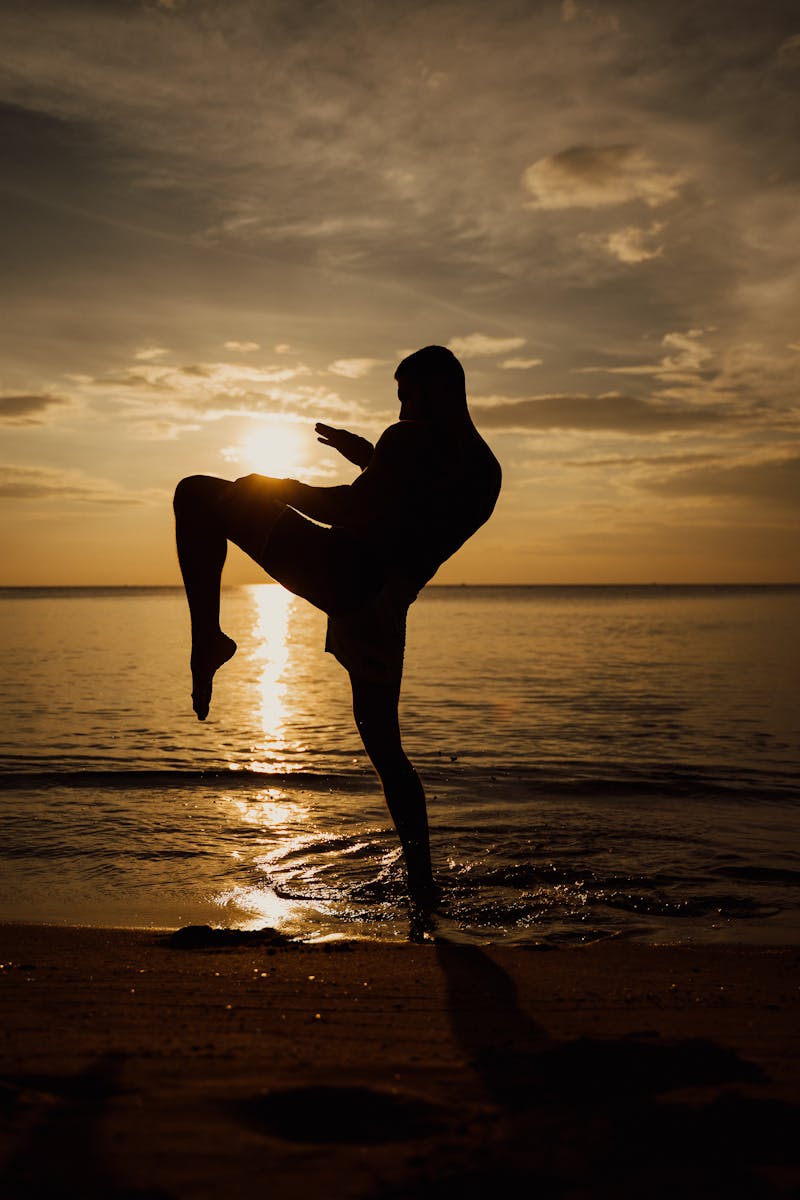 A martial artist practices kickboxing on the beach at sunset, casting a striking silhouette.