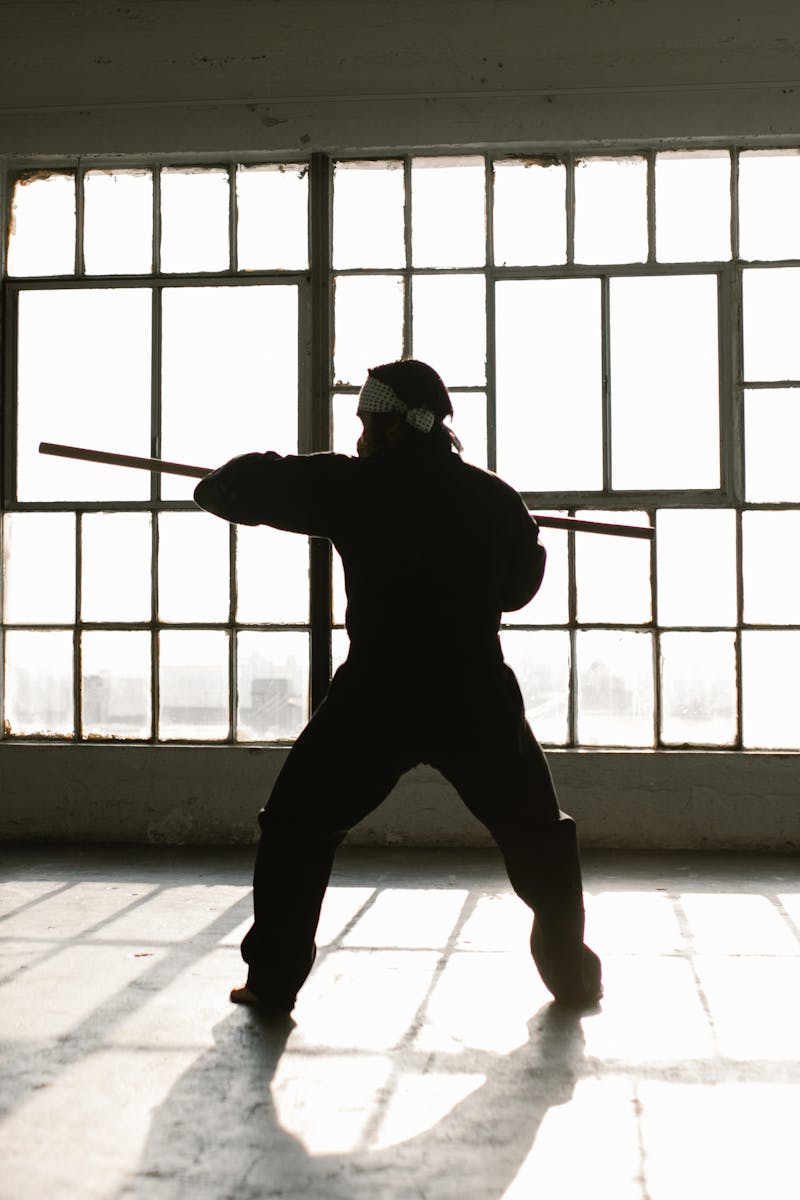 A martial artist practices with a staff in silhouette against large windows.